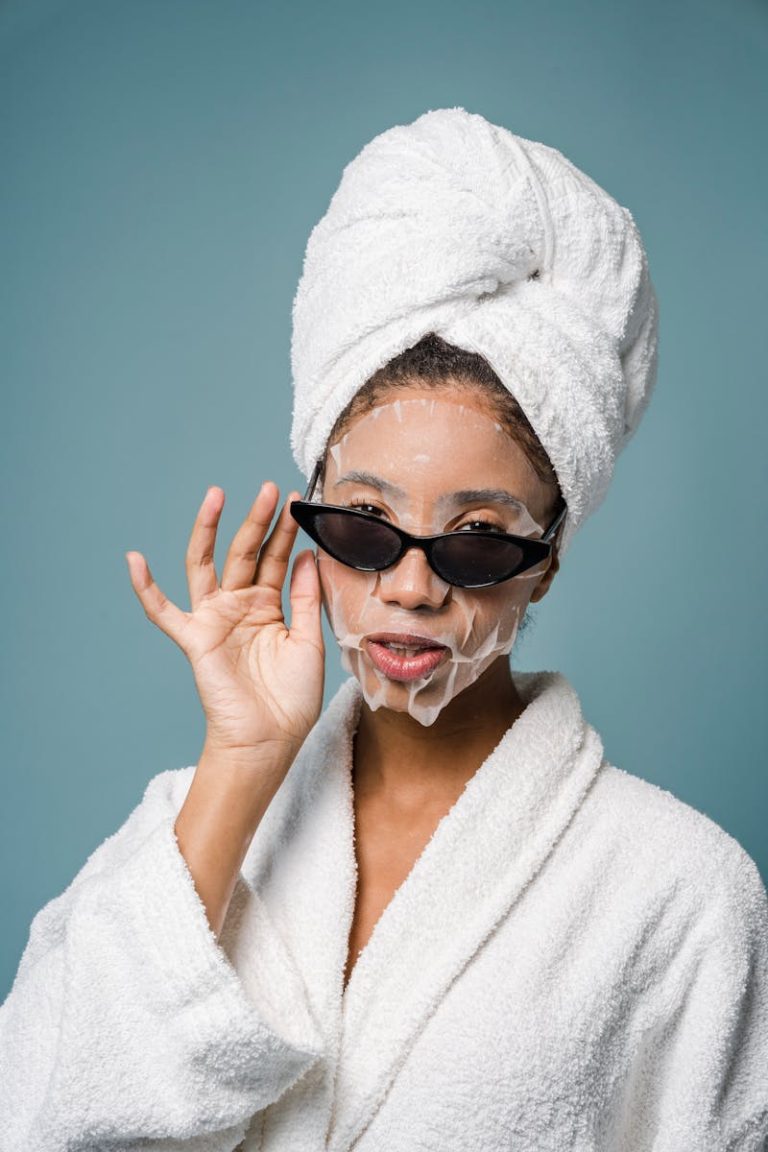 Woman enjoying a skincare routine wearing a towel and sunglasses against a blue background.